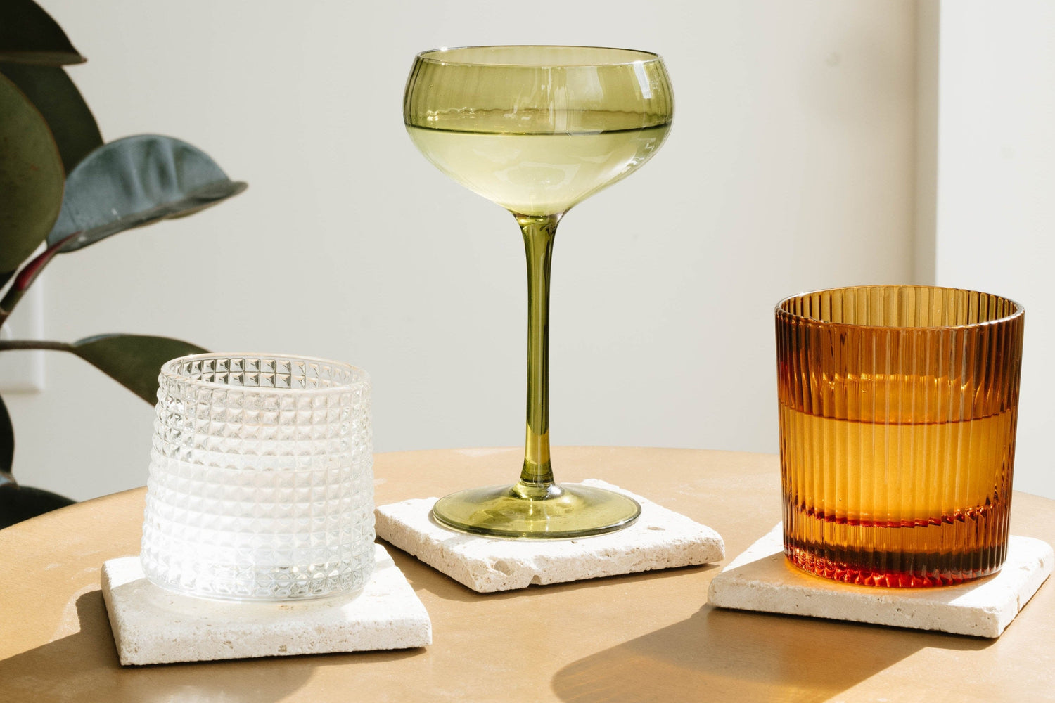 Three textured drinking glasses—one clear, one green, and one amber—sit on square white coasters on a light brown table near a plant.
