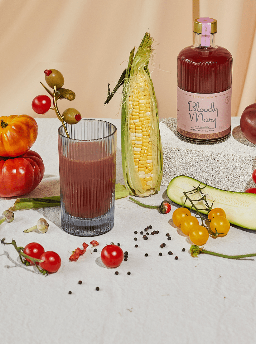 A glass of Bloody Mary with olives, a bottle labeled Bloody Mary, fresh corn, tomatoes, cucumber, cherry tomatoes, and peppercorns arranged on a white cloth.