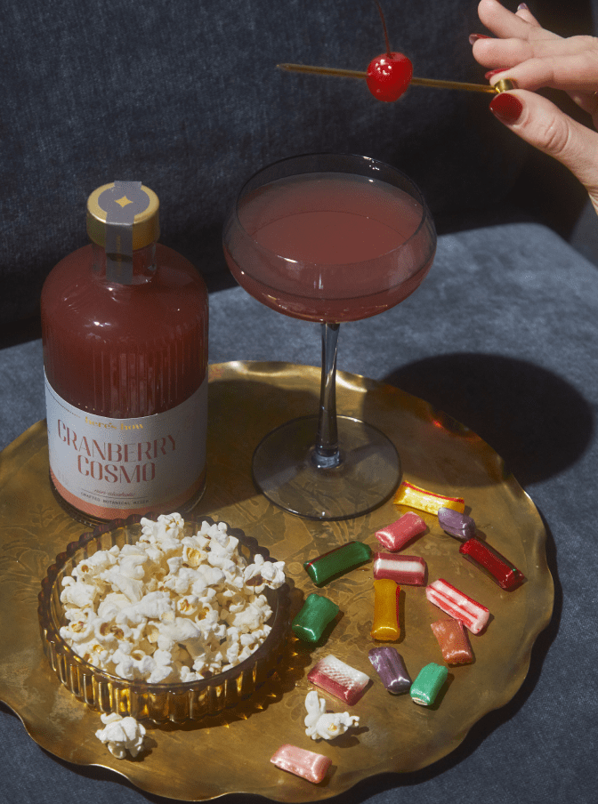 A tray with a glass of cranberry cocktail, a bottle labeled Cranberry Cosmo, a bowl of popcorn, assorted wrapped candies, and a hand holding a cherry above the drink.