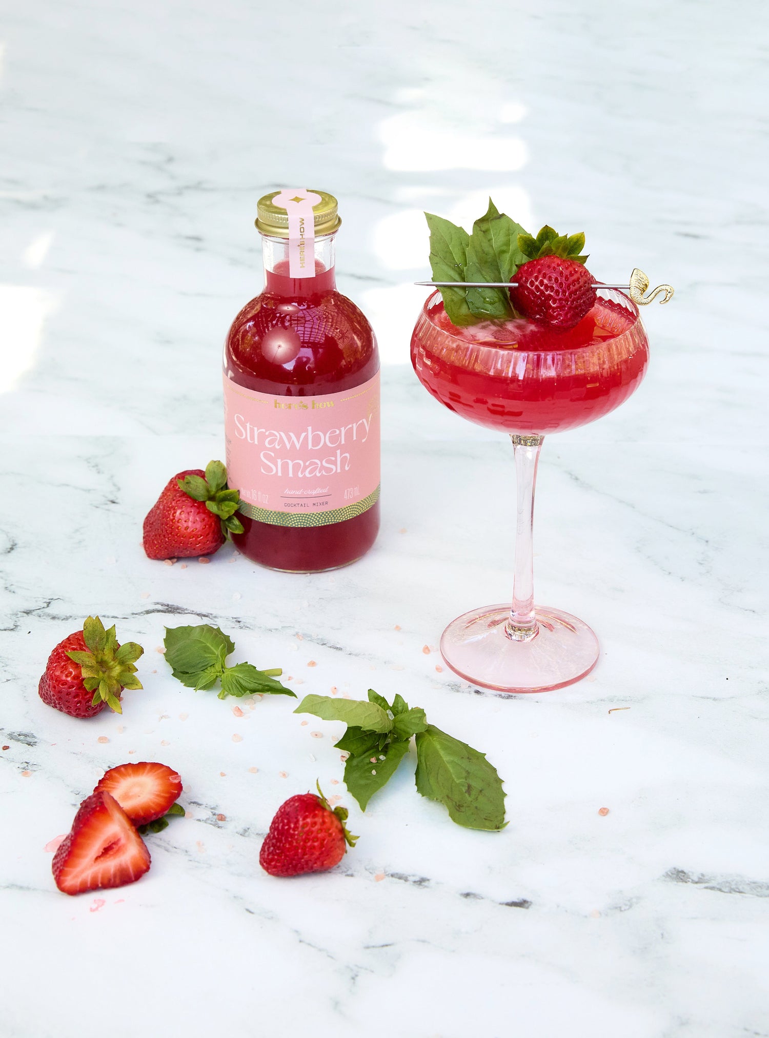 A bottle of Strawberry Smash drink stands next to a cocktail glass filled with a red beverage, garnished with a strawberry and mint, on a marble surface with scattered strawberries and mint leaves.