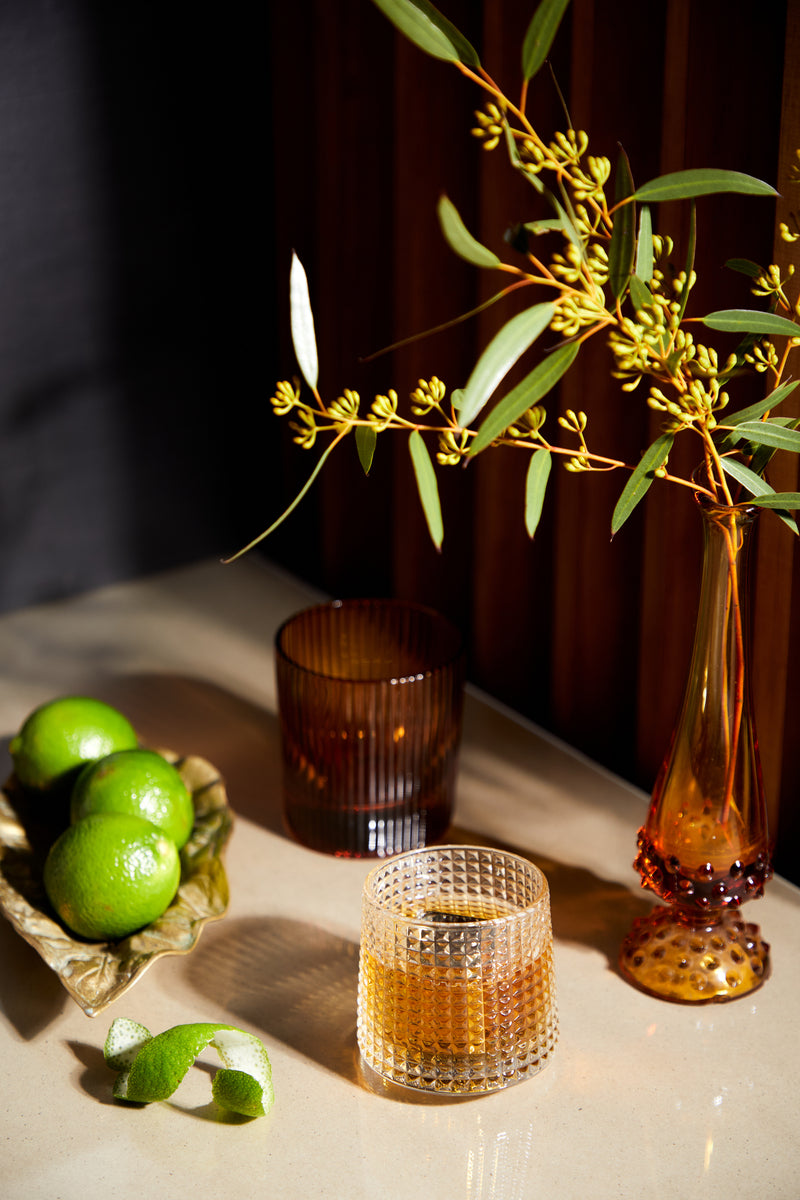The Lola, a fluted stylish tumbler filled with brown liquid, sits on a light countertop next to a ceramic bowl of limes and an amber vase with green foliage.