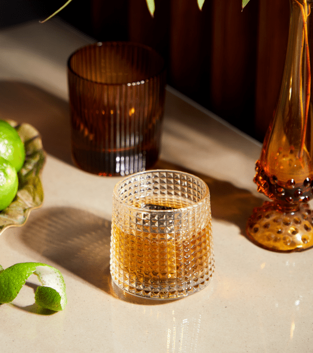 A glass filled with a brown beverage sits on a countertop near a green lime, a lime peel, an amber glass, and an orange decorative vase.