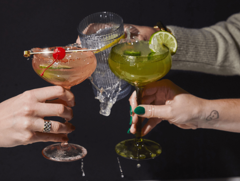 Three hands holding and clinking different cocktails, one pink with a cherry, one clear, and one green with a lime slice; drinks are splashing slightly.