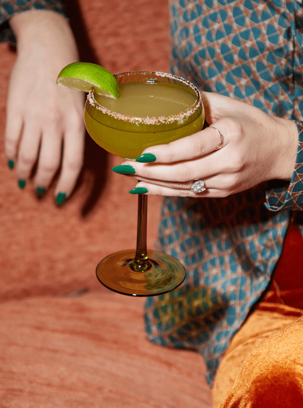 A person with green-painted nails and a ring holds a cocktail glass with a salted rim and lime wedge, sitting on an orange couch.