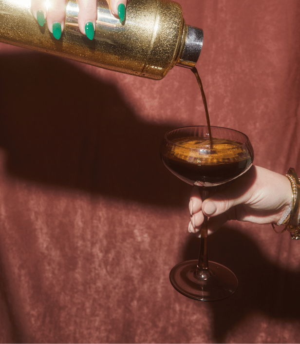 A person pours a dark-colored cocktail from a glittery shaker into a coupe glass, against a brown velvet background.