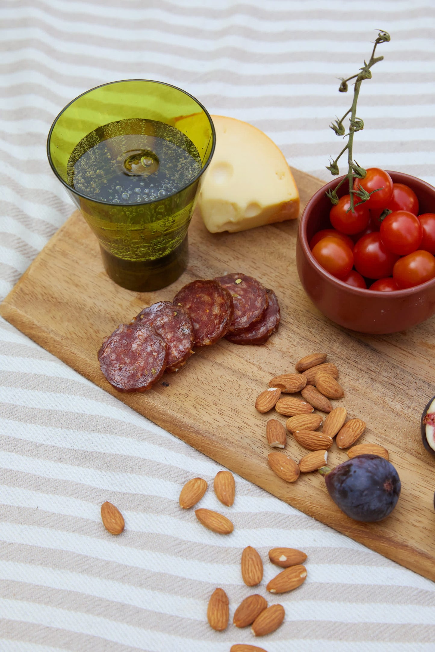 A wooden board with salami, cheese, cherry tomatoes, almonds, and a fig beside The Gilda Tumbler on a striped cloth—perfect for unwinding after sunny beach hours.