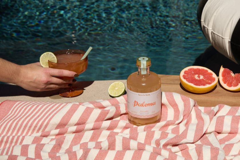 A bottle labeled Paloma sits on a striped towel by the pool at a lively party, with grapefruit, lime, and a hand holding The Rue Margarita Glass garnished with a lime slice.