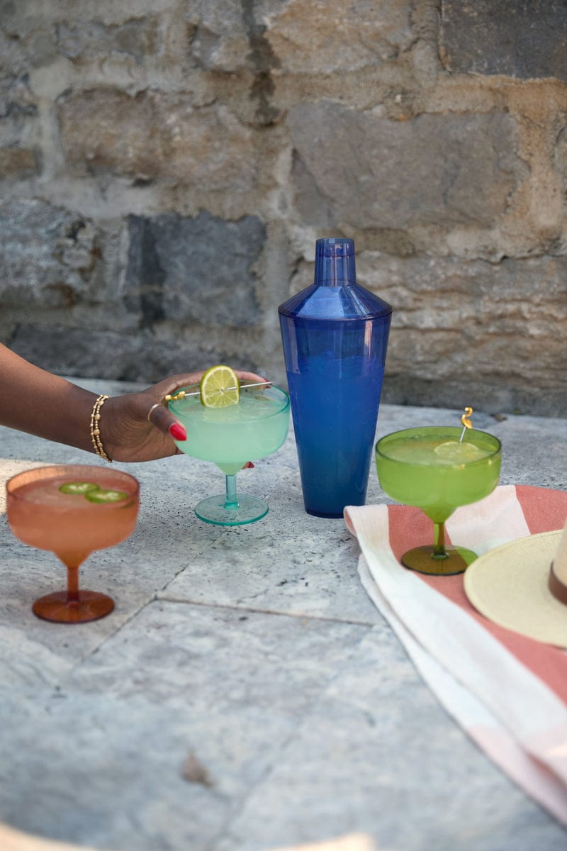 A hand holds a green cocktail glass with lime next to The Penelope Cocktail Shaker in blue, two more drinks, a hat, and a striped cloth on a stone table—ideal for stylish al fresco entertaining.