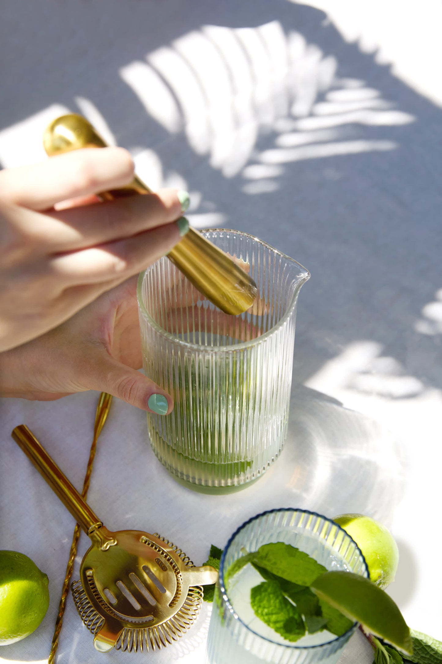 A hand uses The Bertie Barware Set’s gold muddler to crush ingredients in a ribbed glass pitcher. Ideal for cocktail lovers, the set shines in sunlight among a strainer, limes, spoon, and a mint-filled glass.