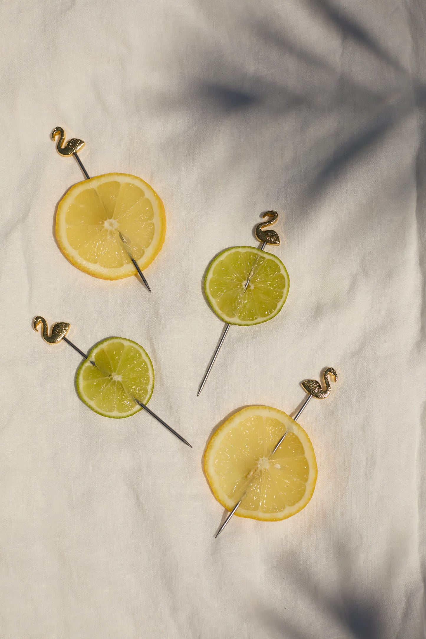 Two lemon and two lime slices, each pierced with The Lily Flamingo Cocktail Picks, arranged on a white fabric background with soft shadows—ideal for adding a tropical touch to your gatherings.