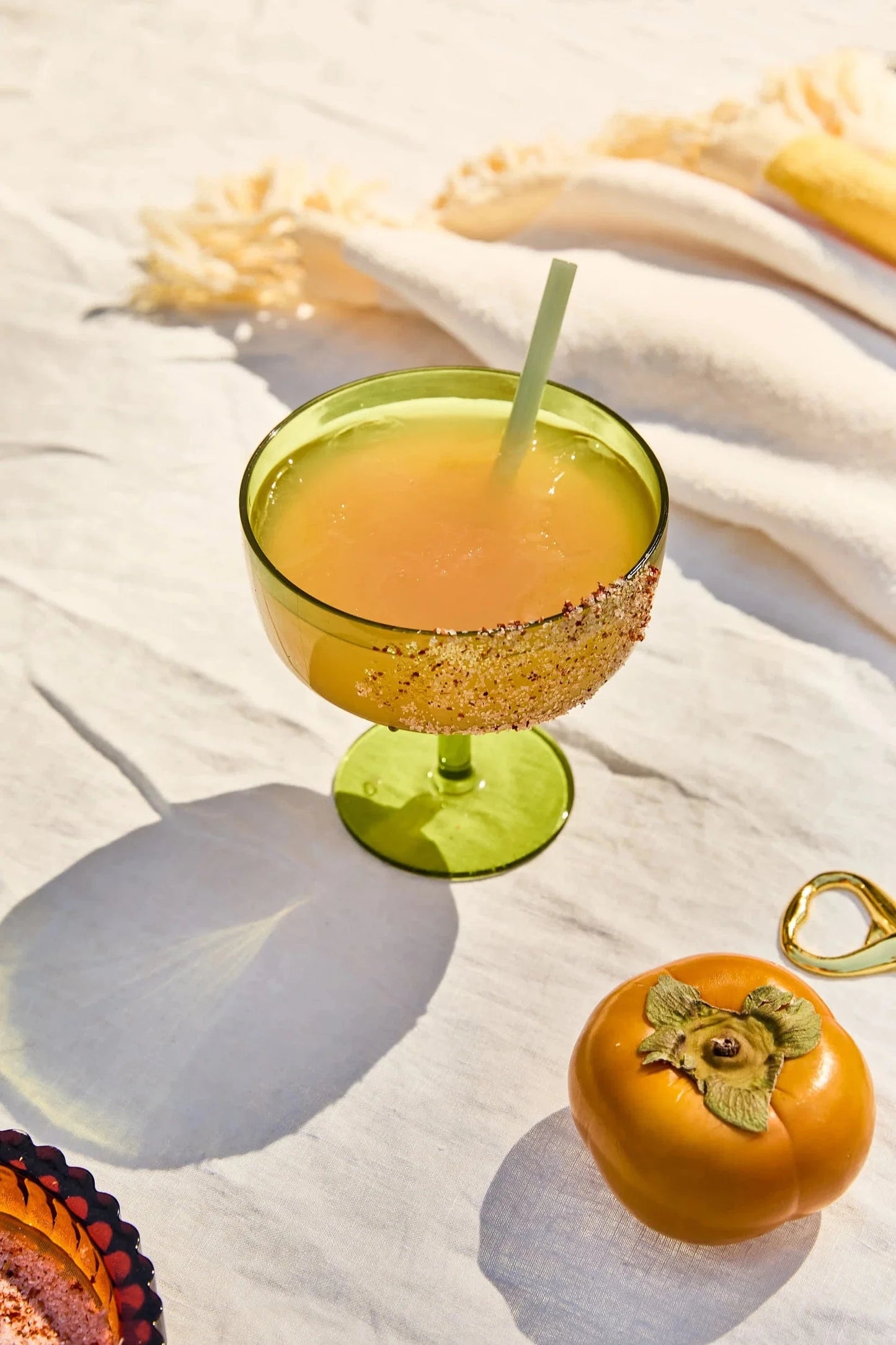 The Margarita Kit: a Rue glass with an orange drink, straw, and seasoned rim sits on white cloth beside a persimmon and gold bottle opener—everything you need for your next margarita mixer.