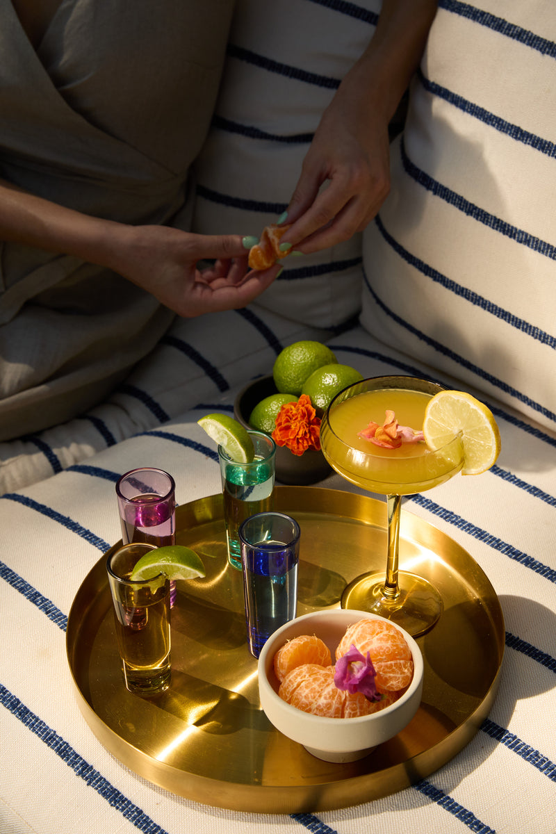 The Gracie Set Of 4 Shot Glasses shine on a gold tray with cocktails and mandarins, creating a festive vibe as someone peels a mandarin in the background.