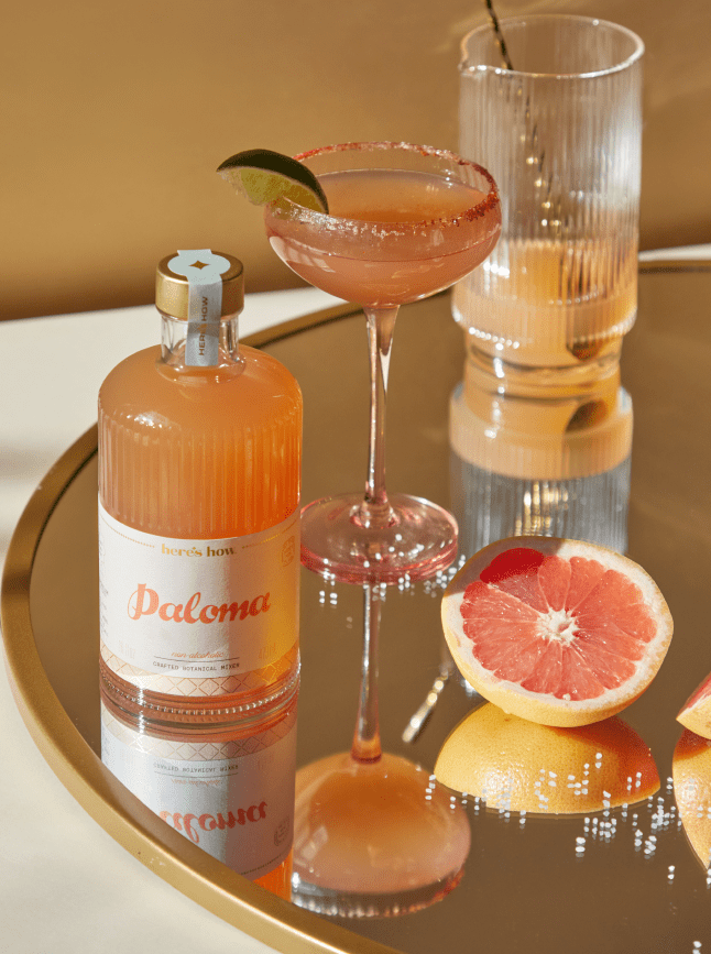 A bottle labeled Paloma, a cocktail in a coupe glass with a lime wedge, a half grapefruit, and a ribbed glass sit on a round mirrored tray.