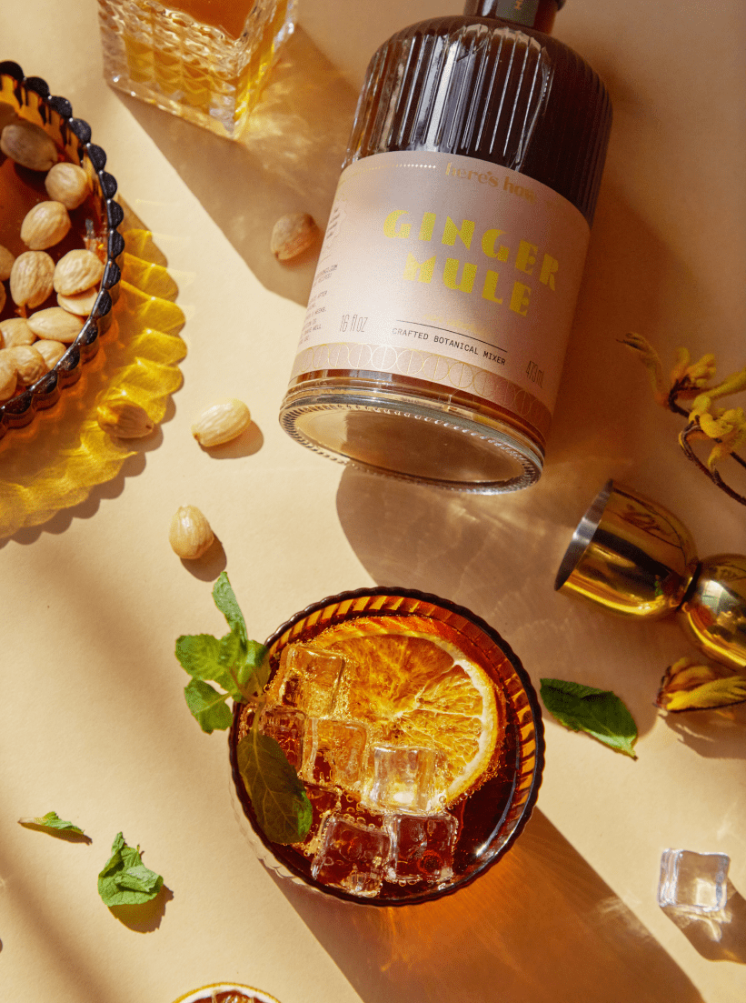 A bottle labeled “Ginger Mule” lies on a table with a cocktail garnished with mint and dried orange, scattered almonds, a gold jigger, and glassware in sunlight.
