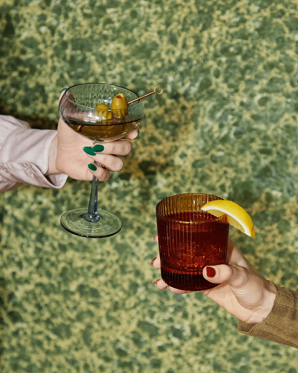 Two hands hold craft cocktails in The Lou coupe glassware: a martini with olives and an amber drink with a lemon wedge, set against a green textured background.