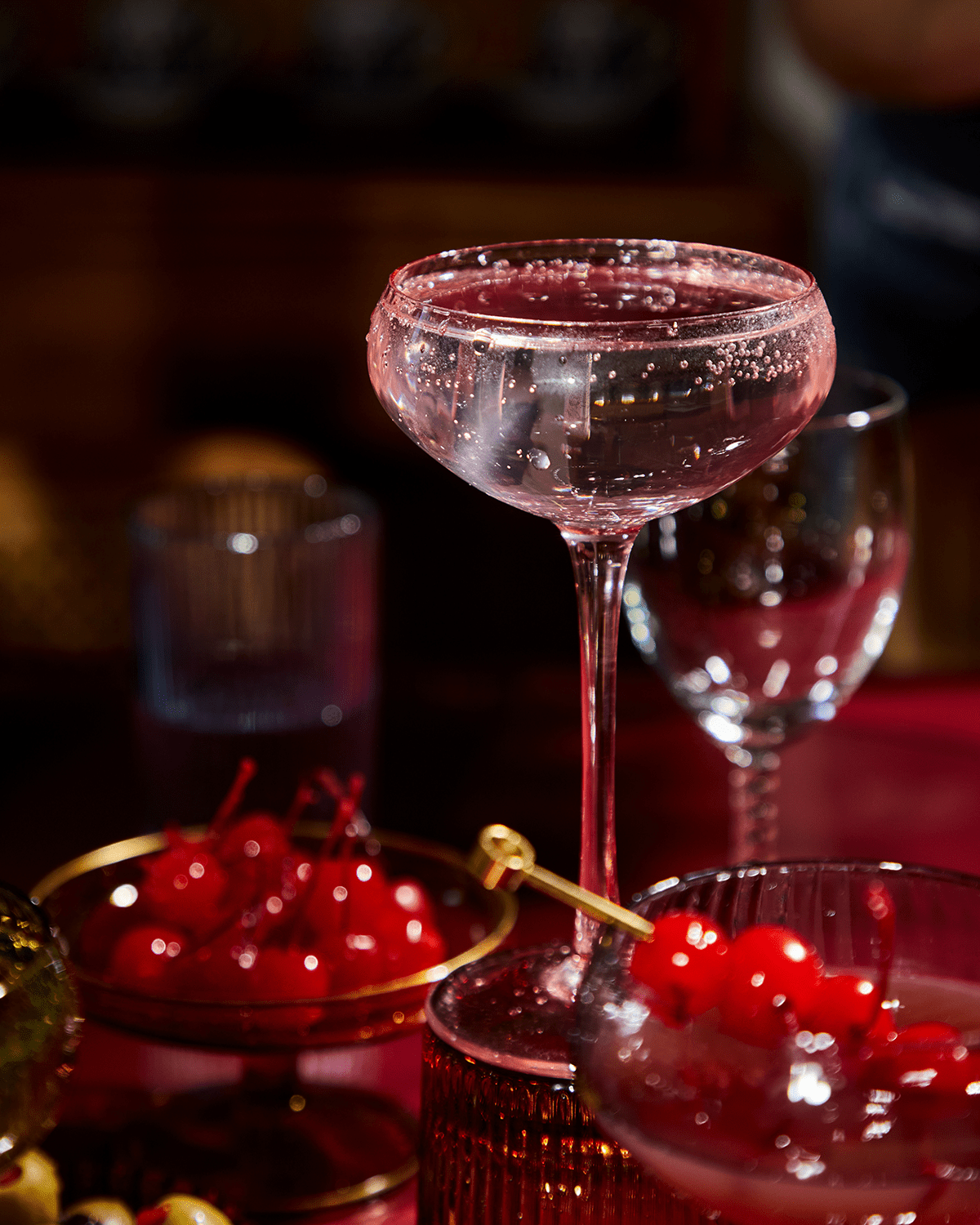 The Lou coupe glassware holds a clear sparkling craft cocktail on a table, with a bowl of maraschino cherries and another Lou glass visible in the background.