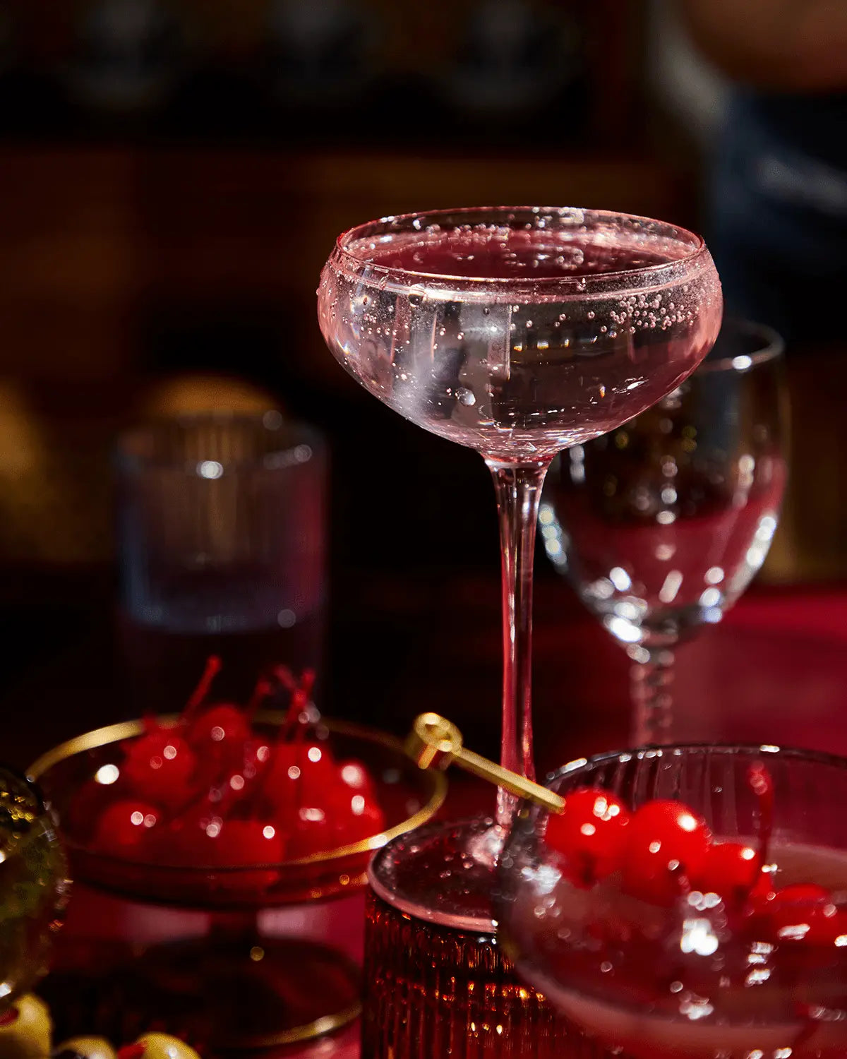 The Lou coupe glassware holds a clear sparkling craft cocktail on a table, with a bowl of maraschino cherries and another Lou glass visible in the background.