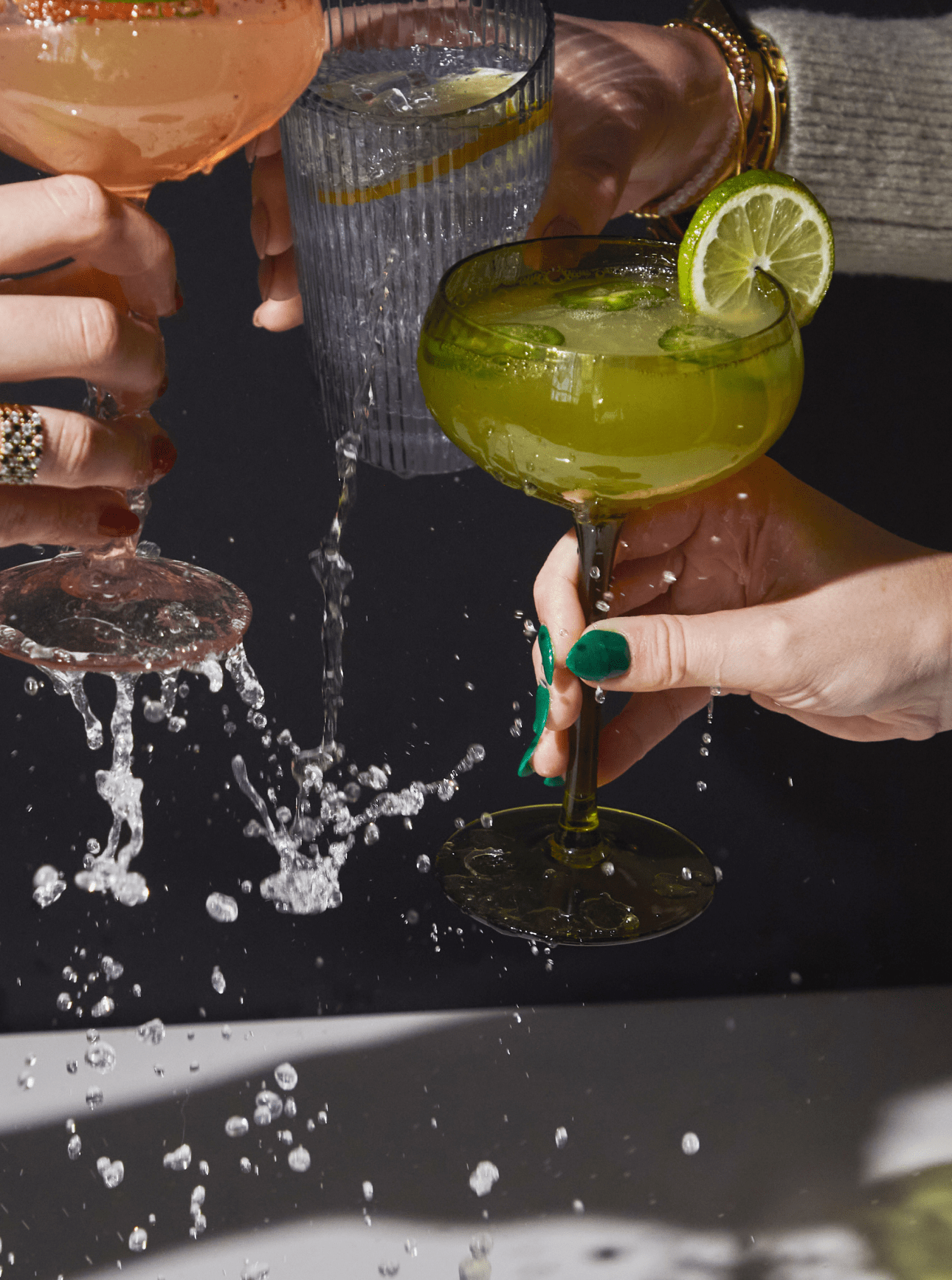 Three hands hold different cocktails as they toast, causing drinks to splash and spill. One glass is garnished with a lime slice.