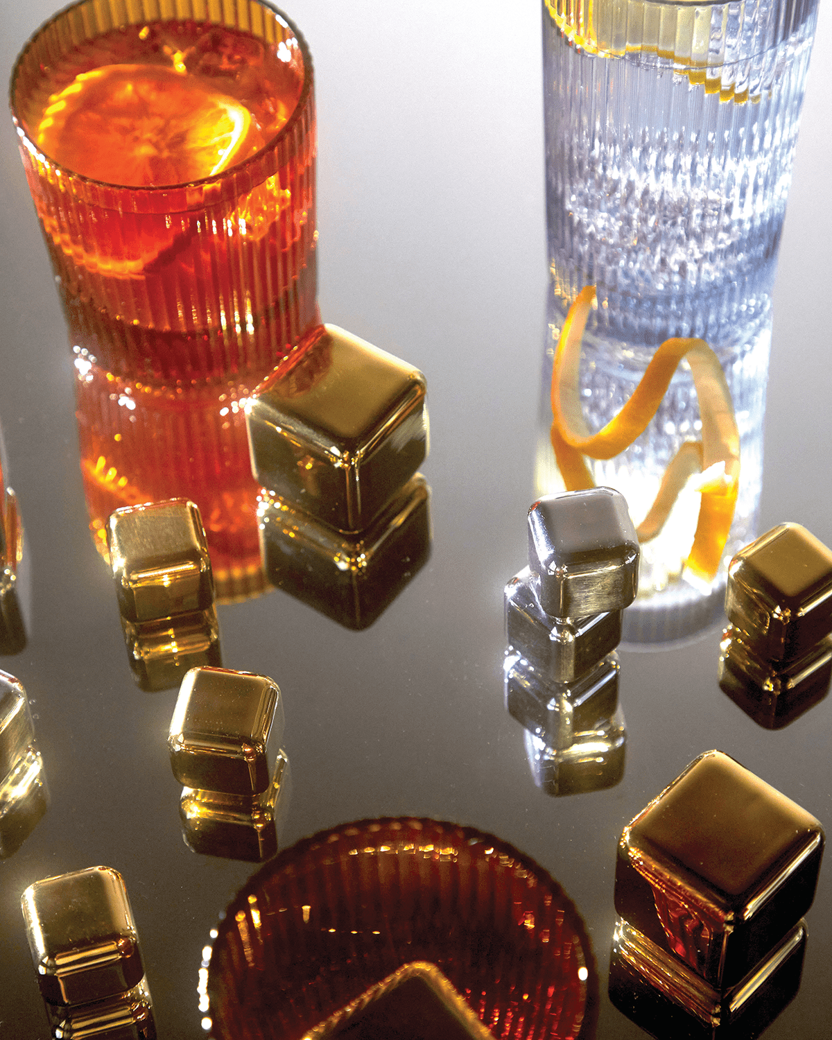 Two Jupiter ribbed glasses with citrus drinks rest on a reflective surface, surrounded by stainless-steel cocktail chillers—reusable, freezable cubes that keep beverages cold without dilution.