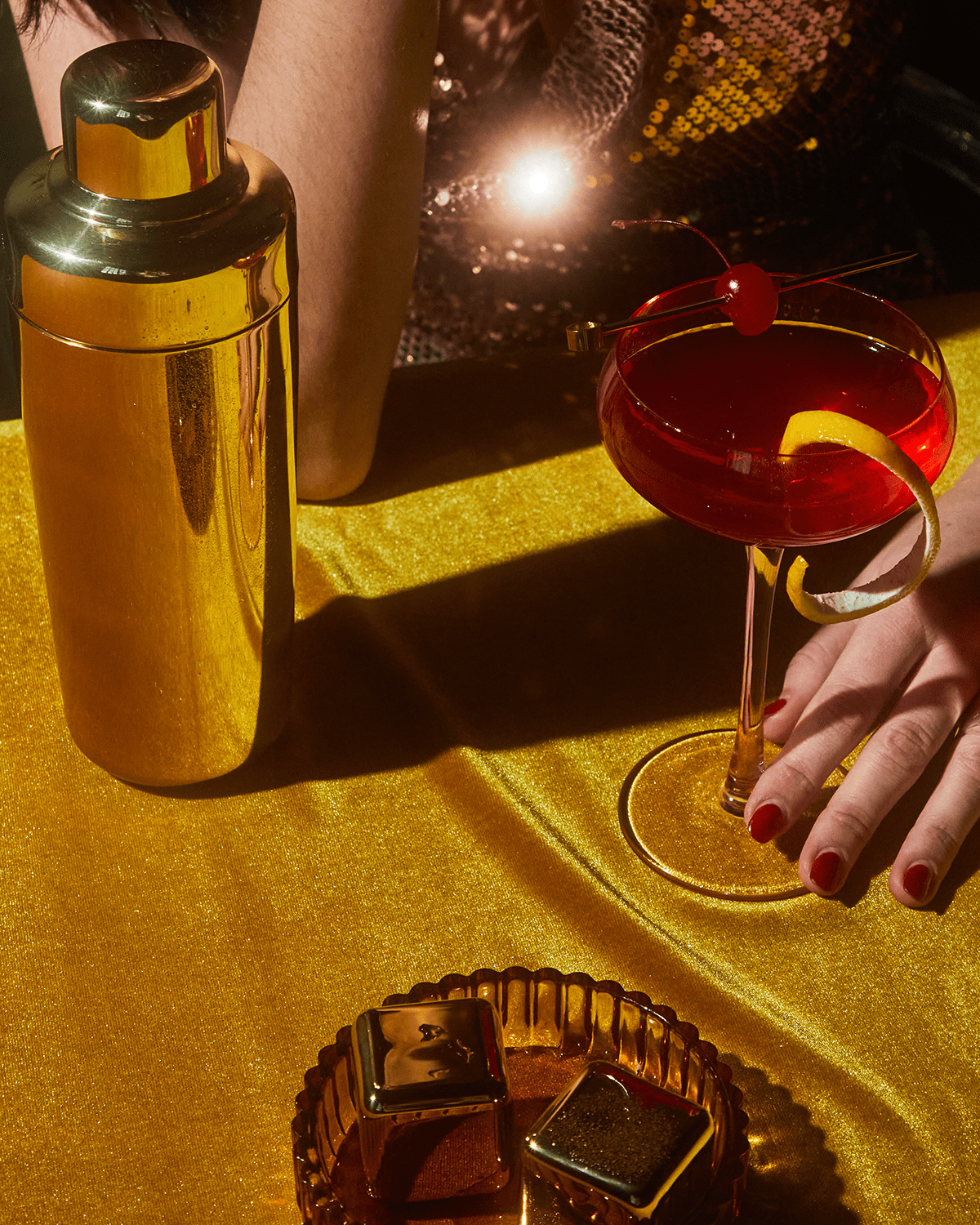 A person with red nail polish holds a cocktail beside The Owen gold shaker and gold-plated cocktail picks on a golden table, with chocolates and sequined clothing in view.