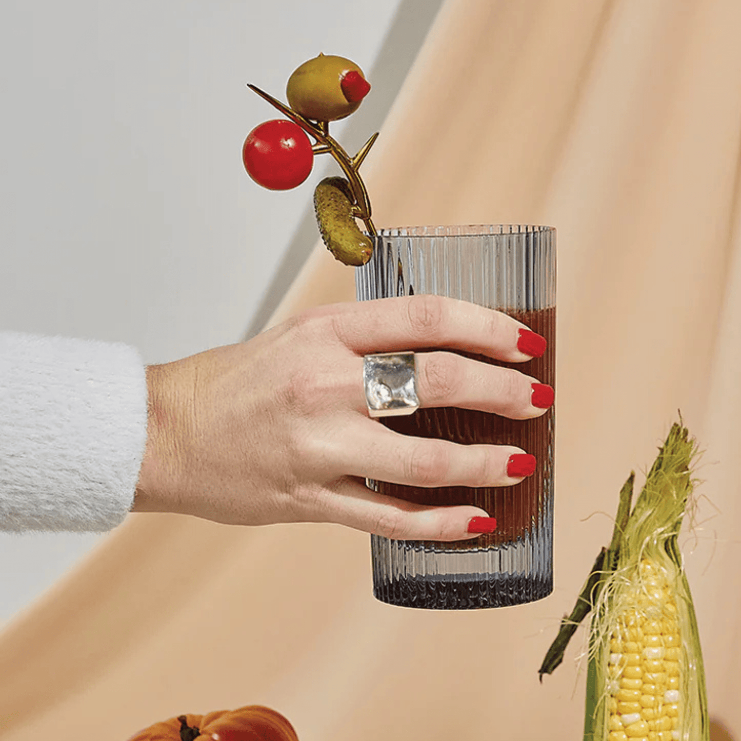 A hand with red nail polish holds a ribbed highball glass of dark liquid, garnished with an olive, tomato, and pickle—perfect for showcasing the Bloody Mary Kit; corn and a pumpkin sit nearby.