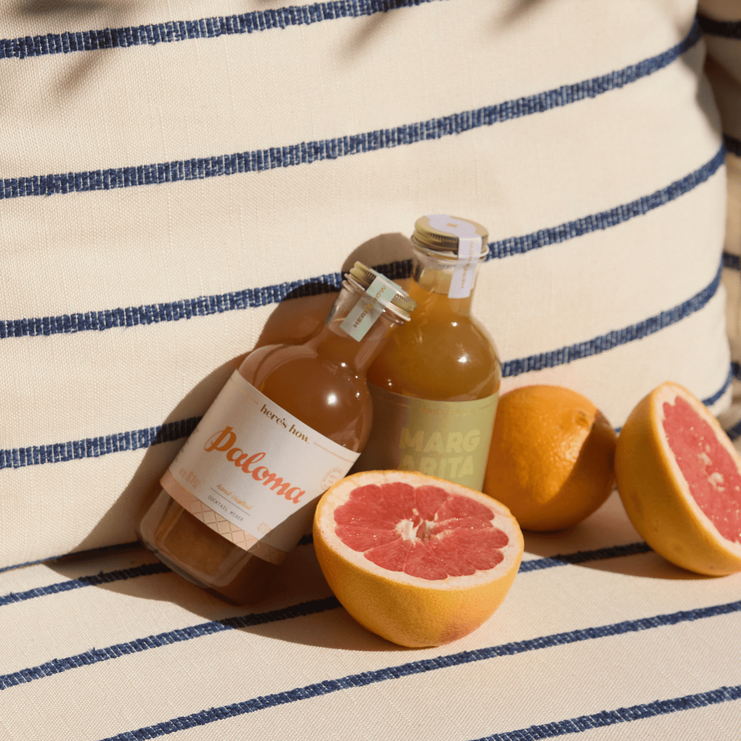 Citrus Duo features two bottled drinks with a halved grapefruit, a whole orange, and another whole grapefruit on a striped cushion—ideal for mixing fresh paloma or margarita tequila cocktails.