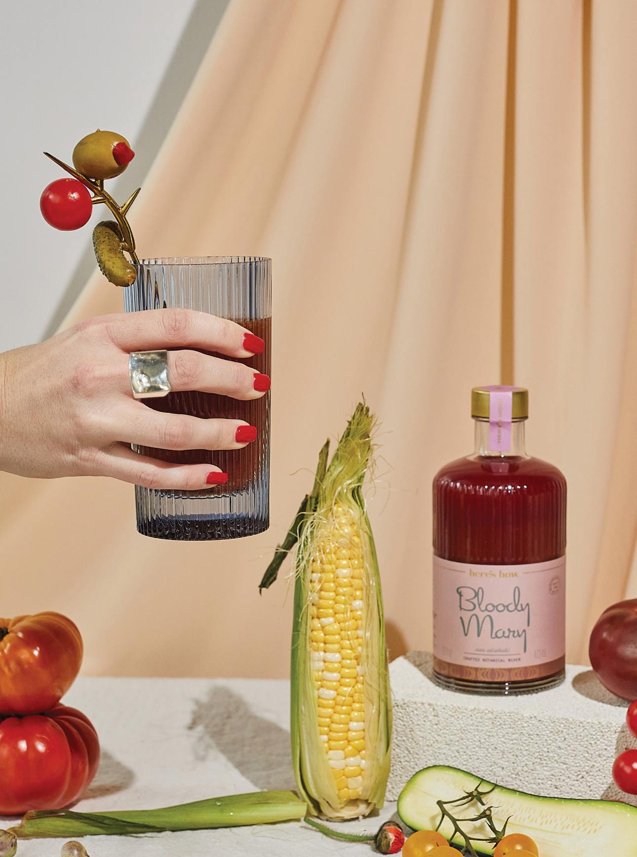 A hand with red nails holds a Bloody Mary garnished with olives and tomatoes; corn on the cob and a bottle labeled Bloody Mary sit nearby, surrounded by fresh vegetables.