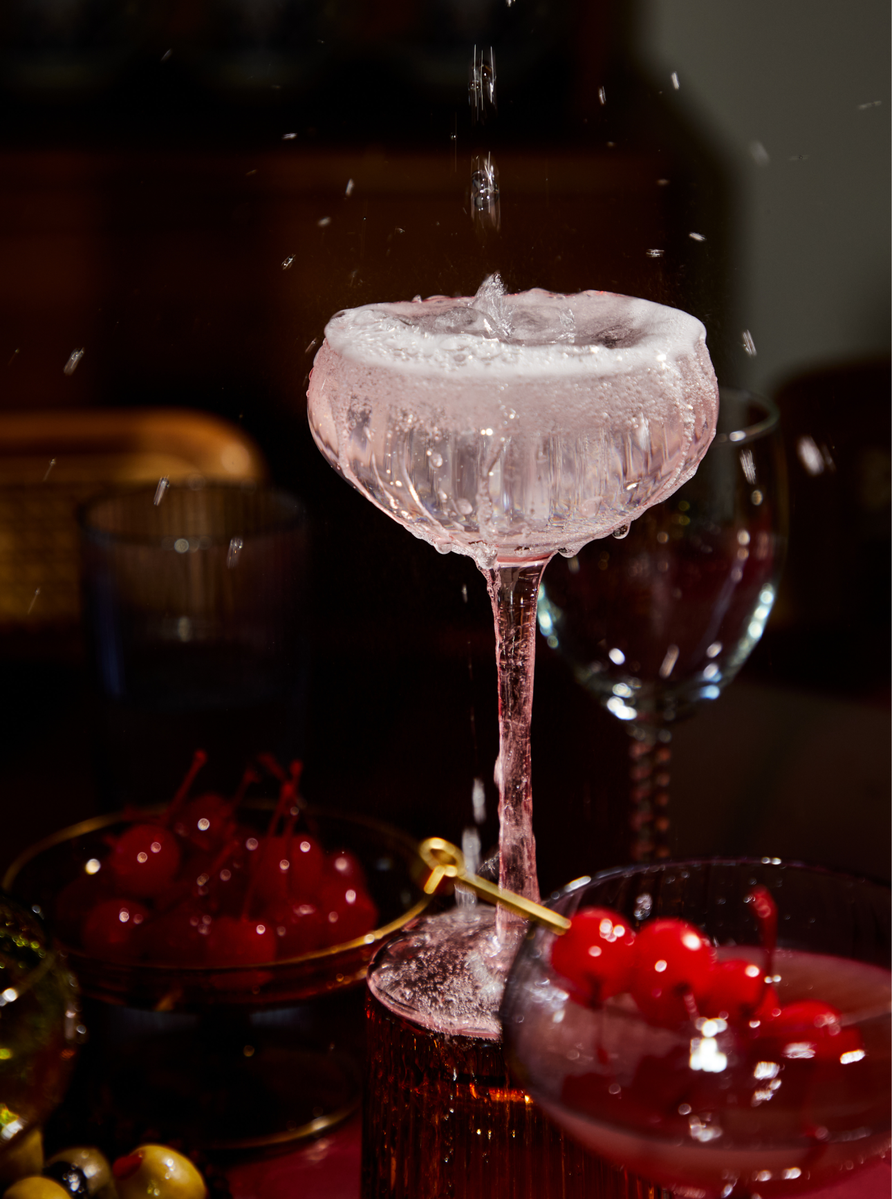 A splash of liquid falls into a coupe glass filled with ice, garnished drinks, cherries, and glassware are visible on the table.
