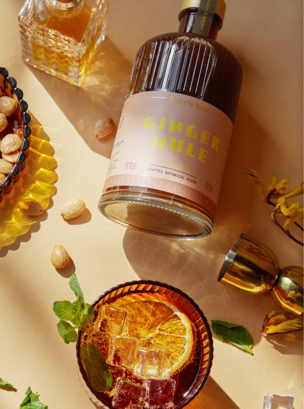 A bottle labeled Ginger Mule lies next to a glass of cocktail with ice, orange slice, and mint, surrounded by nuts, a decanter, and a gold jigger on a beige surface.