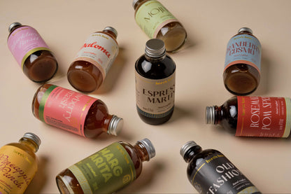 A styled flat-lay arrangement of multiple artisanal whiskey bottles with colorful labels, set against a solid beige background.