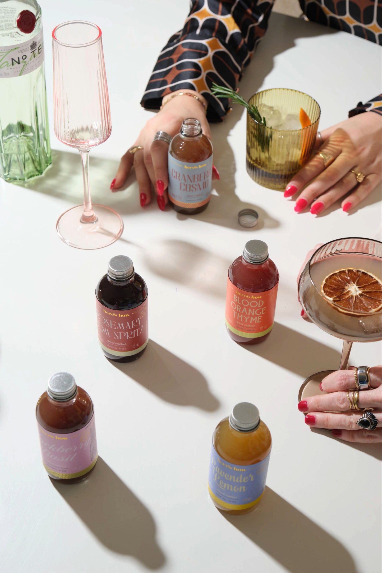 A person arranges the Botanical - Set of 3 Mini Mixers, glasses, and garnishes on a white table; hands with red nails and rings are visible.