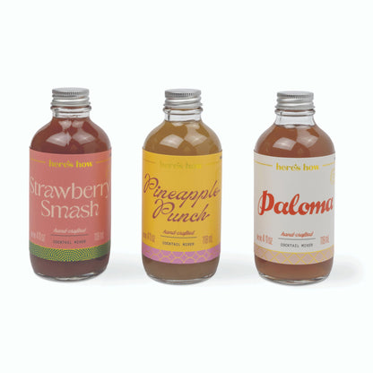 Three small glass bottles of handcrafted cocktail mix with silver caps, labeled Strawberry Smash, Pineapple Punch, and Paloma, arranged side by side on a white background.