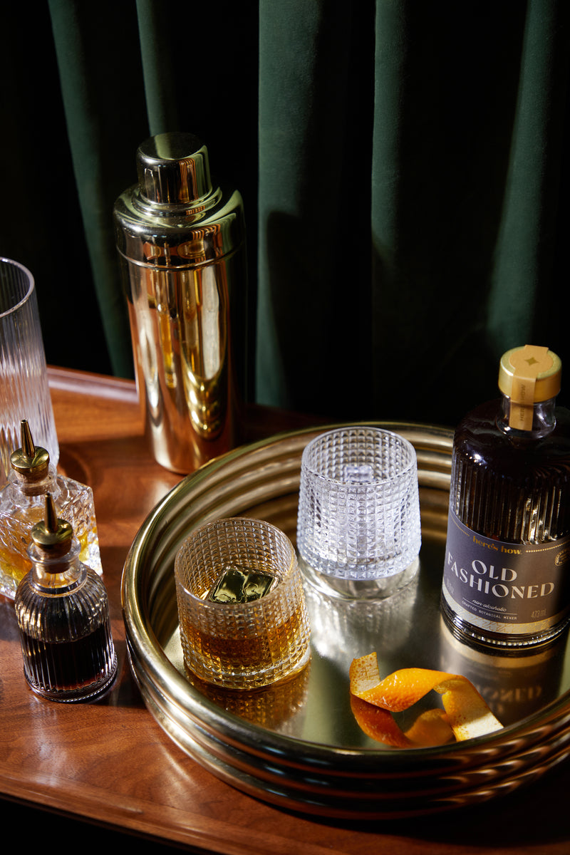 The Lola set features a gold tray with a bottle of Old Fashioned mix, two fluted glasses (one filled, one with ice), a gold cocktail shaker, bitters bottle, and an orange peel garnish.