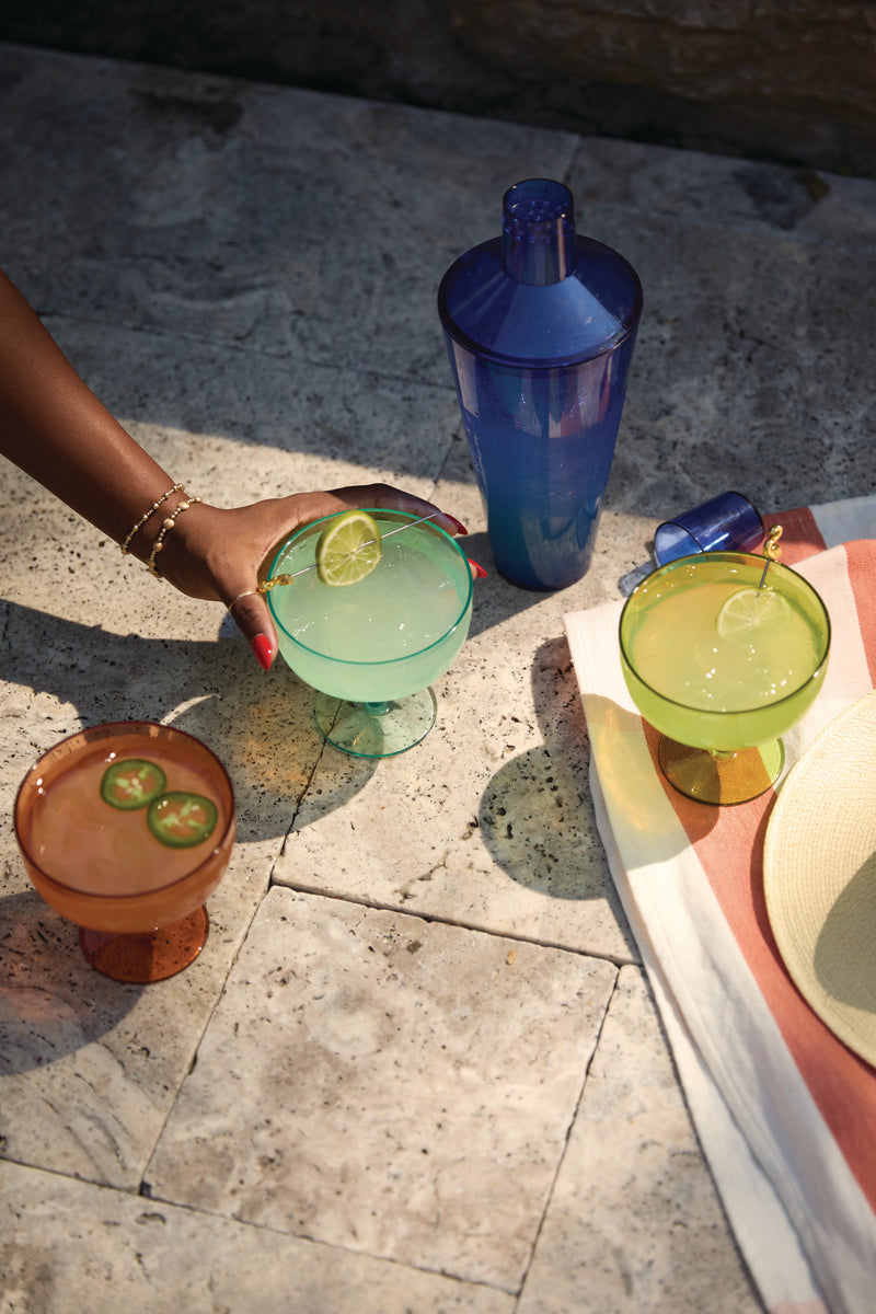 Three vibrant cocktails in The Rue Margarita Glass, garnished and set with a blue shaker on stone, await by a striped towel and hat—a hand reaches for one, making it perfect for sunny days or poolside parties.