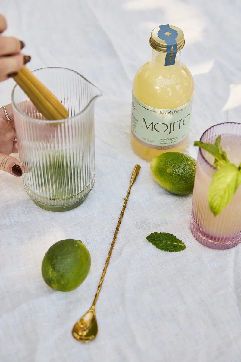 A hand muddles a lime in a glass pitcher next to a bottle of mojito mix, a gold spoon, a whole lime, mint leaves, and a glass with a mint garnish on a white surface.