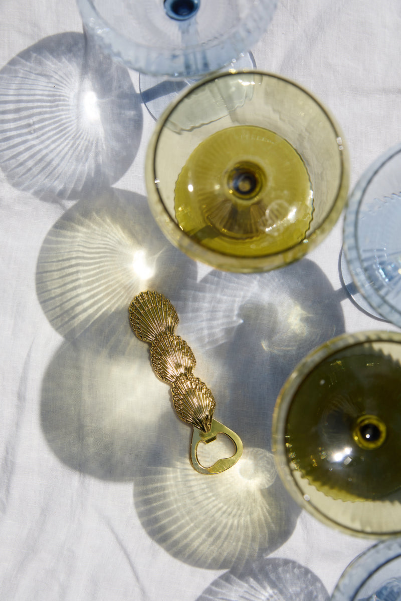 The Theo Shell Bottle Opener, crafted with a gold shell design, rests among translucent wine glasses and casts elegant shadows on a white surface—an ideal gift for beach lovers and cocktail enthusiasts.