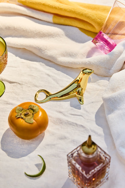 The Blu Seal Bottle Opener, persimmon, sliced lime, pink glass, and glass bottle set on a white cloth with a yellow towel—ocean-inspired essentials for elegant entertaining.