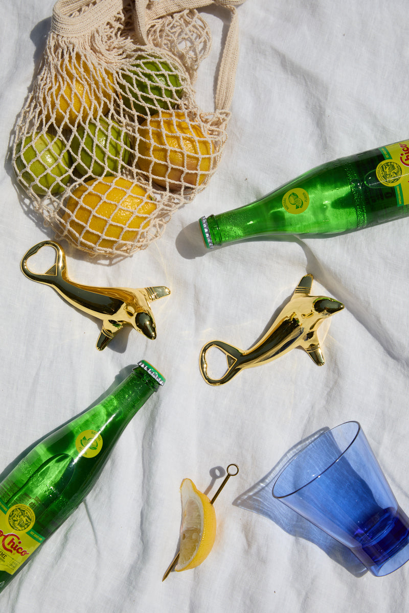 Two green glass Gilda Tumblers, two gold dolphin-shaped bottle openers, a blue glass, a lemon wedge, and a net bag with lemons and limes rest on white cloth—perfect for sun-soaked afternoons and refreshing cocktails.