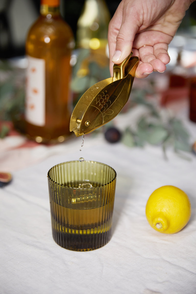A hand squeezes a brass lemon squeezer over a glass of yellow liquid, beside a whole lemon and bottles—the scene highlights The Scout Lowball Glass, perfect for serving your favorite drinks with style.