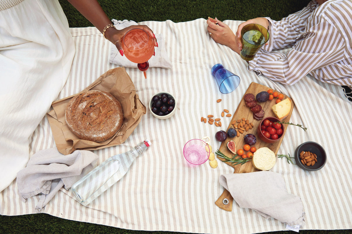 Two people relax on a striped picnic blanket with bread, cheese, grapes, tomatoes, nuts, and drinks served in The Bodie Riviera Glass—ideal for outdoor enjoyment on the grass.
