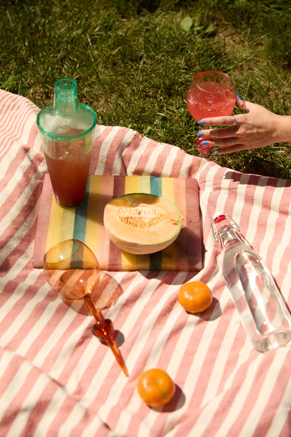 A striped picnic blanket set for outdoor gatherings, featuring The Bodie Riviera Glass with a melon half, two tangerines, a shaker, water bottle, two cocktails, and a hand holding a drink in the top right corner.