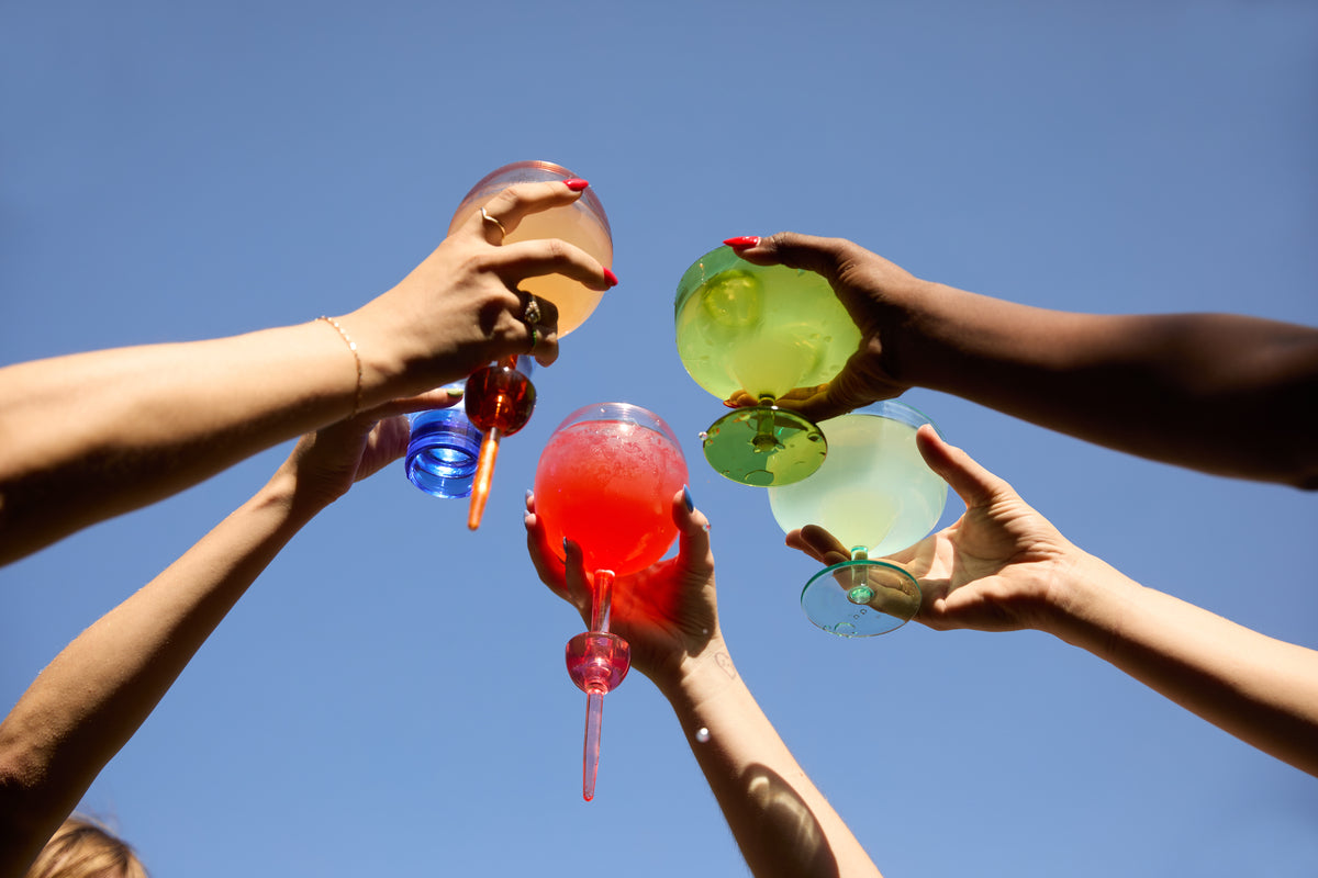 Several hands raise colorful drinks in The Bodie Riviera Glass against a clear blue sky, perfectly capturing the spirit of outdoor enjoyment.