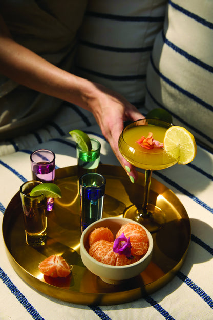 A hand reaches for a cocktail on a gold tray featuring The Gracie Set Of 4 Shot Glasses with lime wedges and a bowl of tangerine segments—an ideal setup for a vibrant party celebration.
