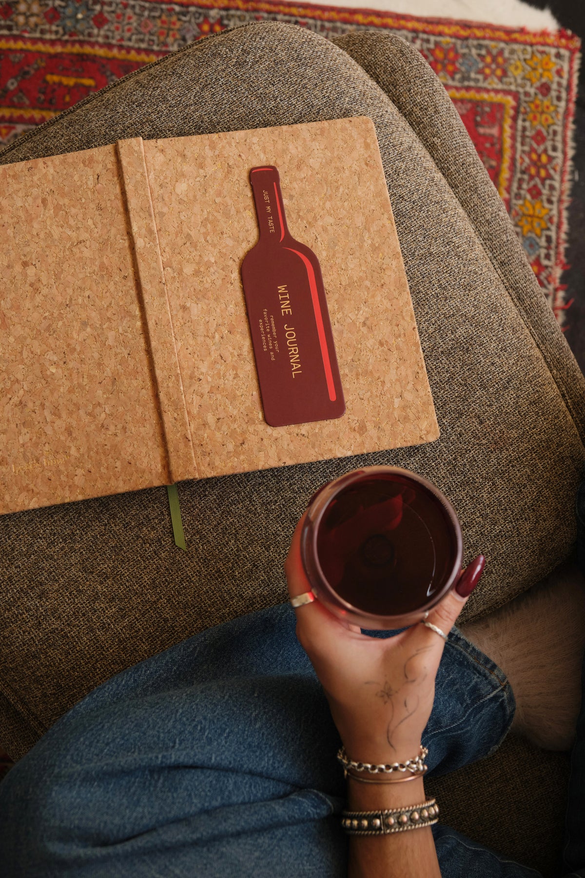 A person holding a glass of red wine sits on a couch near their Wine Journal - Cork Wine Bottle, jotting down tasting notes with the journal’s wine bottle-shaped bookmark resting on top.