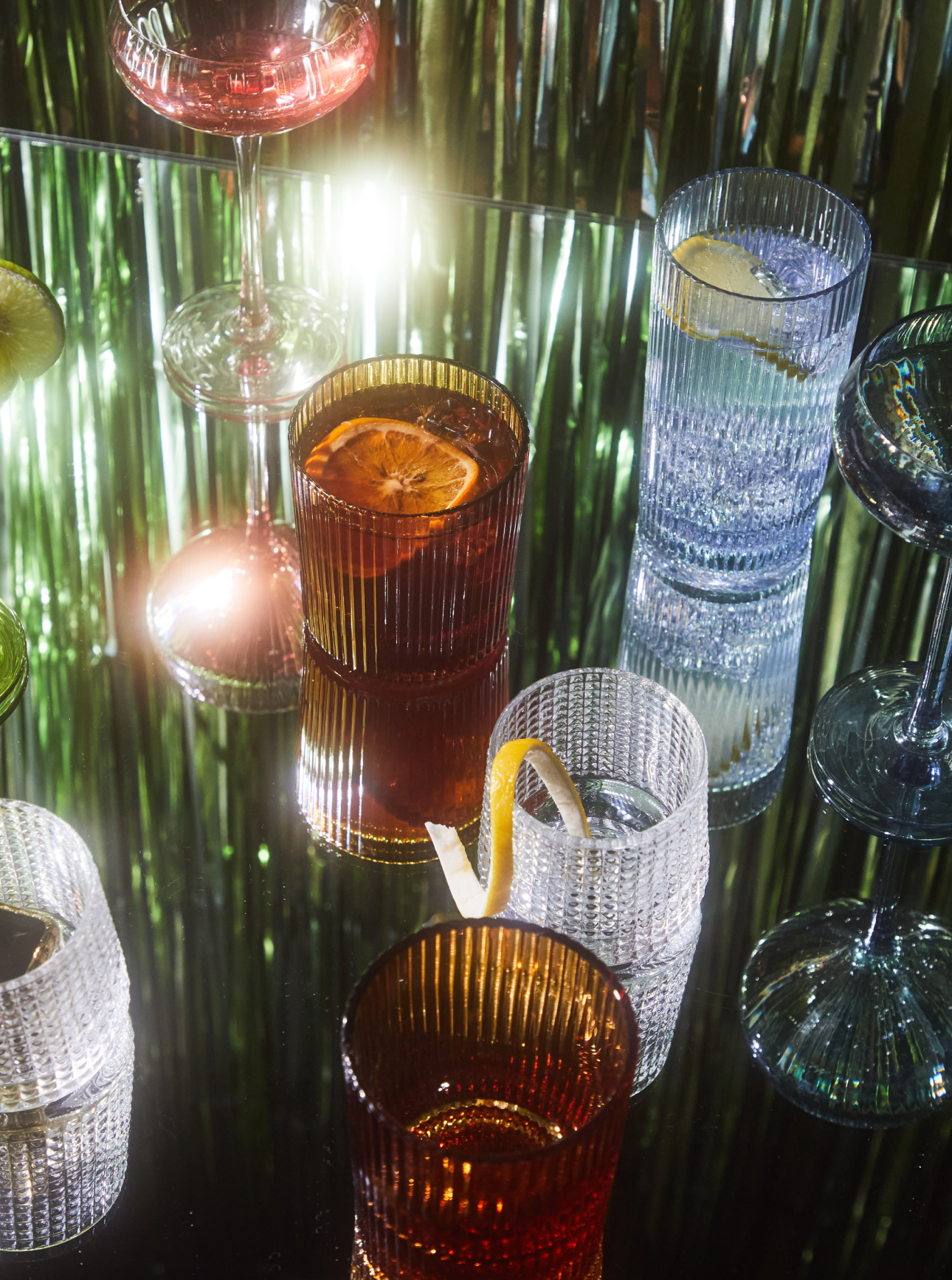 Various glassware with cocktails and garnishes are arranged on a reflective surface with metallic green and silver streamers in the background.