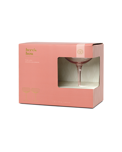A pink box labeled Heres How features The Lou coupe glassware set, with one craft cocktail coupe partly visible through a front transparent window.