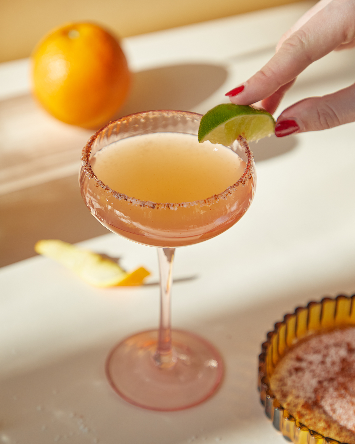 A hand with red nail polish places a lime wedge on the rim of The Lou, served in a salted coupe glass; an orange and orange peel sit nearby.