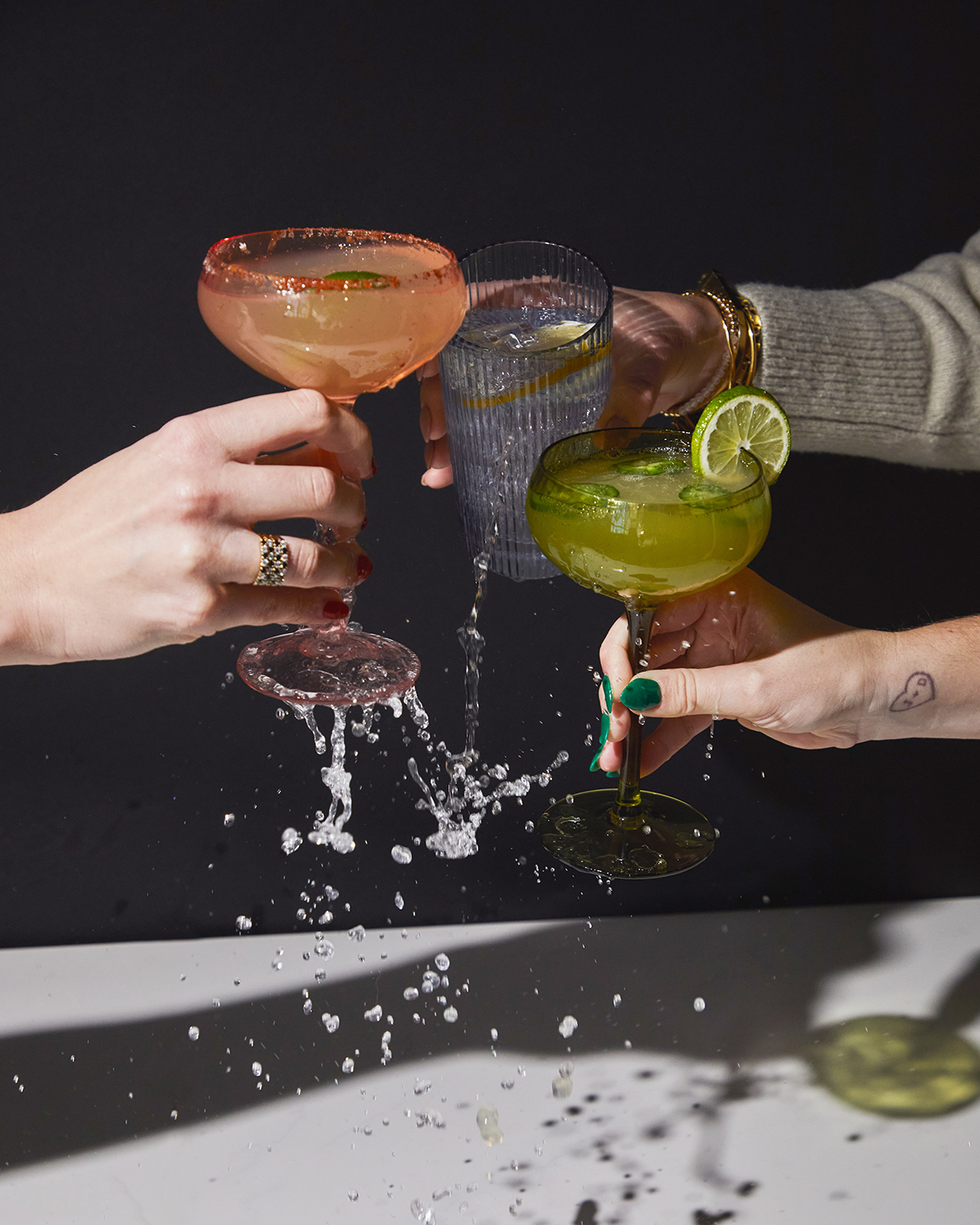 Three hands raise craft cocktails in a celebratory toast, splashing drinks onto a white surface against a black background. One hand holds The Lou soda lime glass filled with water, while the others have margaritas in coupe glasses.