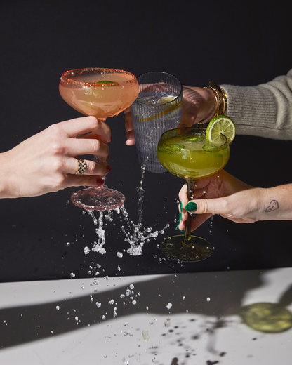 Three hands raise craft cocktails in a celebratory toast, splashing drinks onto a white surface against a black background. One hand holds The Lou soda lime glass filled with water, while the others have margaritas in coupe glasses.