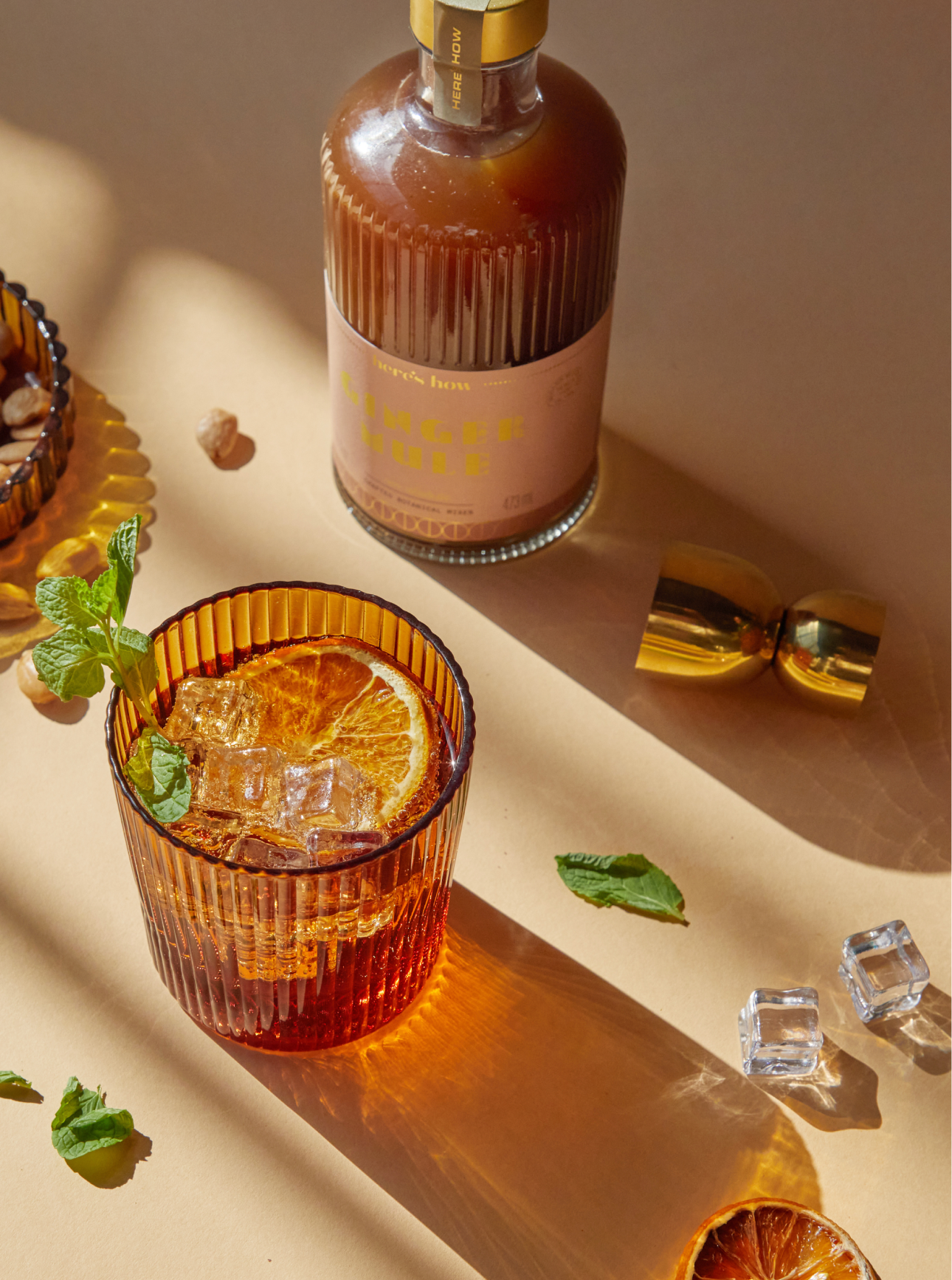 A glass of iced cocktail garnished with a dried orange slice and mint sits next to a brown bottle, golden jigger, ice cubes, and mint leaves on a beige surface.